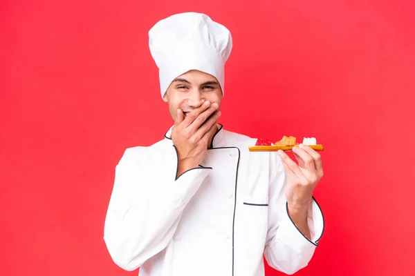赤い背景に孤立した刺身を持つ若い白人料理人幸せと笑みを浮かべて手で口をカバー — ストック写真