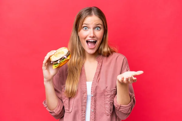 Młoda Kaukaska Kobieta Trzyma Hamburgera Czerwonym Tle Wstrząśniętym Wyrazem Twarzy — Zdjęcie stockowe