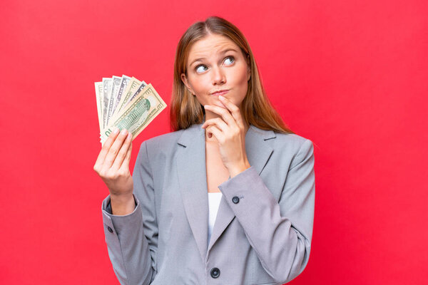 Young business caucasian woman holding money isolated on red background having doubts while looking up