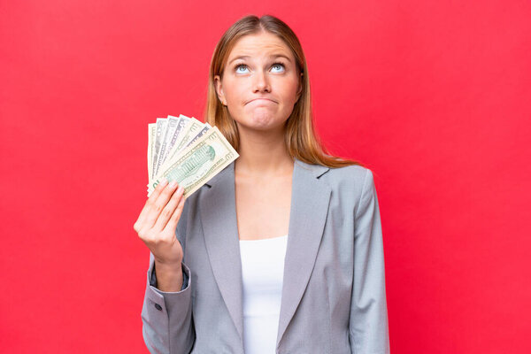 Young business caucasian woman holding money isolated on red background and looking up