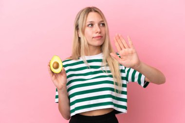 Pembe arka planda avokado tutan beyaz bir kadın dur hareketi yapıyor ve hayal kırıklığına uğruyor.