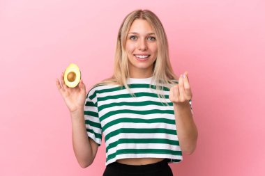 Avokado tutan beyaz bir kadın pembe arka planda izole edilmiş para hareketi yapıyor.