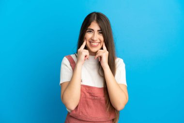 Mavi arka planda izole edilmiş genç beyaz kadın mutlu ve hoş bir ifadeyle gülümsüyor.
