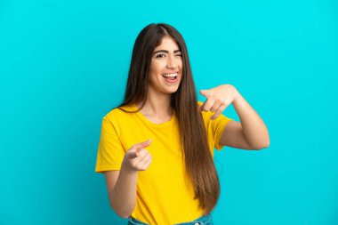 Mavi arka planda izole edilmiş genç beyaz kadın ön tarafı işaret ediyor ve gülümsüyor.