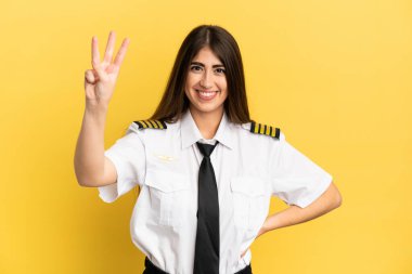 Uçak pilotu sarı arka planda izole edilmiş mutlu ve parmaklarıyla üçe kadar sayıyor