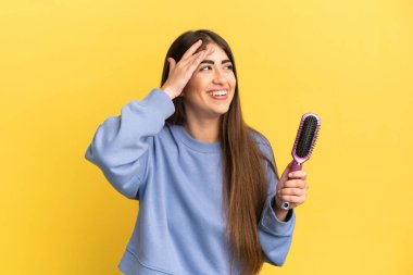 Mavi arka planda saç fırçası tutan beyaz bir kadın çok fazla gülümsüyor.