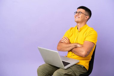 Genç adam dizüstü bilgisayarlı bir sandalyede oturmuş gülümserken yukarı bakıyor.