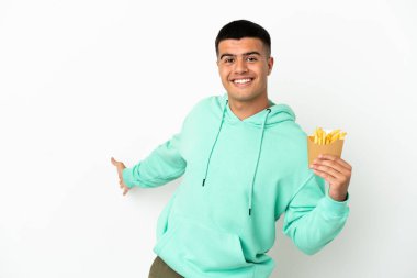 Genç ve yakışıklı bir adam izole edilmiş beyaz arka planda patates kızartması tutuyor. Davet etmek için ellerini yana uzatıyor.