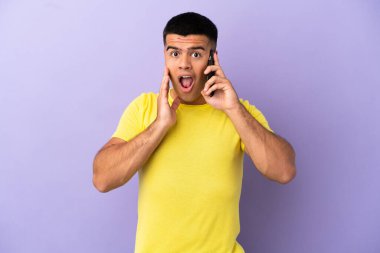 İzole edilmiş mor arka planda, şaşırmış ve şok olmuş bir yüz ifadesiyle cep telefonu kullanan genç bir adam.