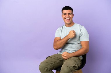 Young man sitting on a chair over isolated background celebrating a victory