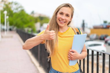 Açık havada genç sarışın bir bayan. Başparmakları havada. Çünkü iyi bir şey oldu.