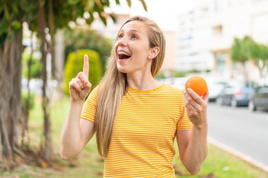 Açık havada portakal tutan genç sarışın kadın parmağını kaldırarak çözümü anlamaya çalışıyor.