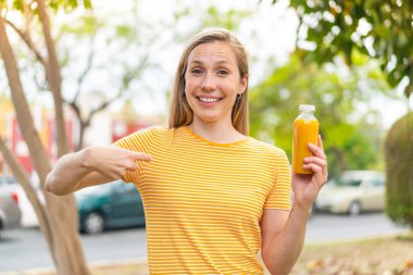 Açık havada elinde portakal suyu tutan sarışın genç bir kadın. Sürpriz bir yüz ifadesi var.