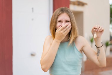 Açık havada genç sarışın bir kadın elinde mutlu bir ifadeyle evin anahtarlarını tutuyor.