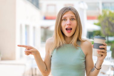 Açık havada kahve taşıyan genç sarışın kadın şaşırdı ve parmağını yana çevirdi.