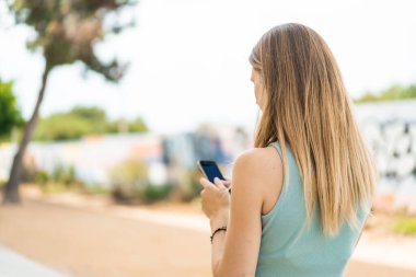 Arka tarafta cep telefonu kullanan genç sarışın kadın.