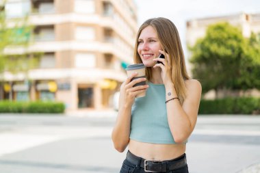 Açık havada cep telefonu kullanan ve elinde mutlu bir ifadeyle kahve tutan genç sarışın kadın.