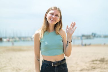 Dışarıda genç sarışın bir kadın, mutlu bir ifadeyle selamlıyor.