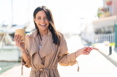 Açık havada kahve taşıyan genç bir kadın açık havada şaşkın şaşkın ve parmağıyla bir tarafı işaret ediyor.
