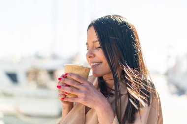 Açık havada elinde neşeli bir ifadeyle kahve taşıyan genç bir kadın.