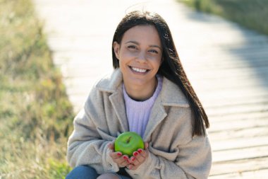 Açık havada elinde mutlu bir ifadeyle elma tutan genç bir kadın.