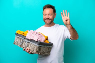 Mavi arka planda izole edilmiş çamaşır sepetini tutan orta yaşlı bir kadın mutlu ve parmaklarıyla dört tane sayıyor.