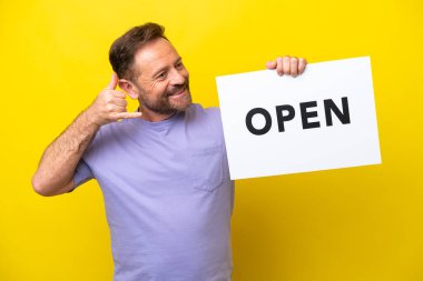 Orta yaşlı beyaz bir adam sarı arka planda izole edilmiş. Elinde açık yazılı bir levha var ve telefon hareketi yapıyor.