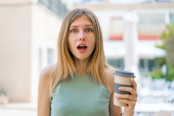 若いですブロンド女性持っています取り上げコーヒーで屋外で驚きとショックを受けた顔の表情 — ストック写真