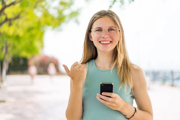 Giovane Donna Bionda All Aperto Utilizzando Telefono Cellulare Indicando Laterale — Foto Stock