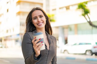 Genç bir kadın elinde kahveyle dışarıda mutlu bir ifadeyle bekliyor.