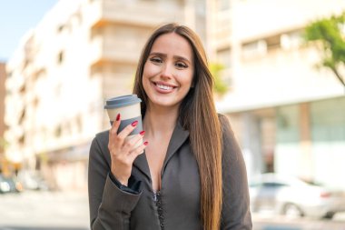 Açık havada elinde neşeli bir ifadeyle kahve taşıyan genç bir kadın.