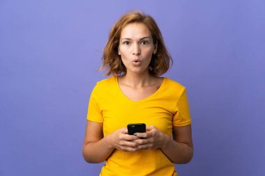 Genç Gürcü kadın mor arka planda izole edilmiş kameraya bakarken şaşırmış bir ifadeyle cep telefonunu kullanıyor.