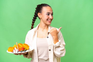 İzole edilmiş arka planda waffle tutan genç bir Arap kadın bir ürünü sunmak için kenarı işaret ediyor.