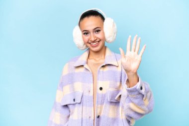 Mavi arka planda izole edilmiş kış pantolonu giyen genç Arap kadın parmaklarıyla beşi sayıyor.