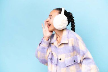 Mavi arka planda izole edilmiş, ağzı sonuna kadar açık bir şekilde bağıran, kışın takılan genç Arap kadın.