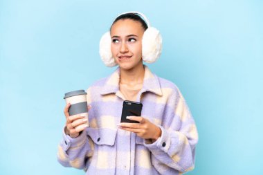 Mavi arka planda izole edilmiş kışlık kuklalar takan genç bir Arap kadın elinde kahve ve bir şeyler düşünürken bir cep telefonu tutuyor.