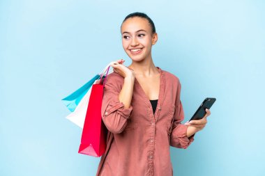 Mavi arka planda izole edilmiş genç bir Arap kadın elinde alışveriş çantaları ve bir cep telefonu tutuyor.