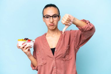 Genç bir Arap kadın elinde bir kâse meyve tutuyor mavi arka planda izole edilmiş başparmağını negatif bir ifadeyle gösteriyor.