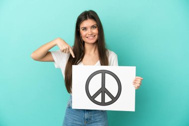 Kafkasyalı genç bir kadın mavi arka planda izole edilmiş. Elinde barış sembolü olan bir levha var.