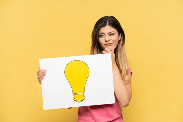 Young Russian Girl Isolated Yellow Background Holding Placard Bulb Icon — Stock Photo, Image
