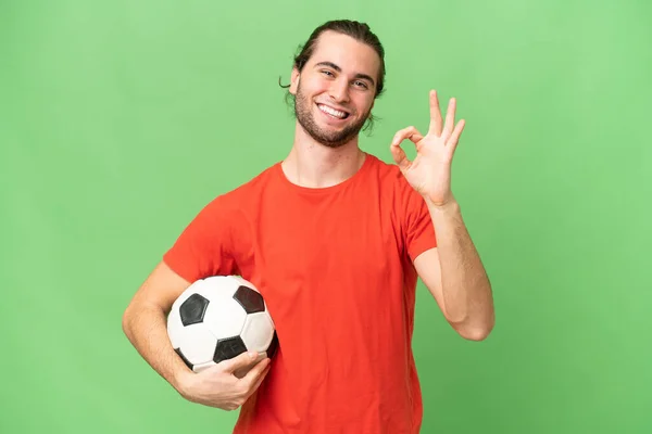 若いですハンサムな男隔離上の緑のクロマ背景とともにサッカーボールと作りますOkサイン — ストック写真