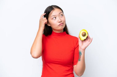 Beyaz arka planda avokado tutan Asyalı genç bir kadının şüpheleri ve kafa karıştırıcı bir yüz ifadesi var.