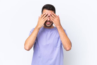 Beyaz arka planda izole edilmiş beyaz tenli genç adam gözlerini elleriyle kapatıyor.