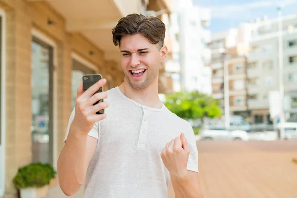 Mladý Pohledný Muž Pomocí Mobilního Telefonu Dělá Vítězství Gesto — Stock fotografie