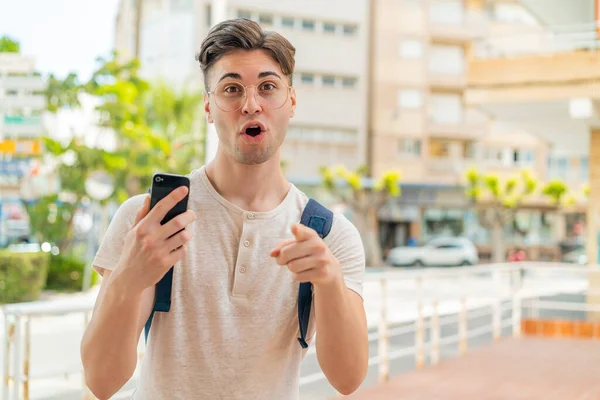 Mladý Pohledný Muž Pomocí Mobilního Telefonu Venku Překvapen Ukazuje Dopředu — Stock fotografie