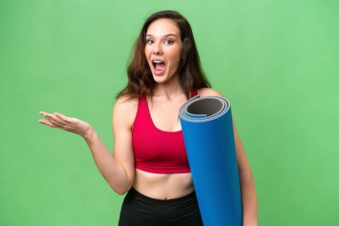 Genç sporcu kadın yoga derslerine giderken elinde yalıtılmış bir arka plan ve şok olmuş yüz ifadesi ile bir minder tutuyor.