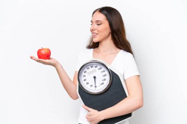 Beyaz arka planda izole edilmiş genç beyaz bir kadın elmaya bakarken bir tartı makinesi tutuyor.