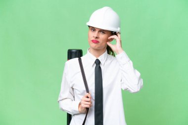 Young architect woman with helmet and holding blueprints over isolated background having doubts