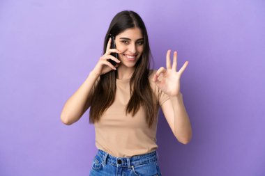 Mor arka planda izole edilmiş cep telefonu kullanan beyaz bir kadın parmaklarıyla işaret veriyor.