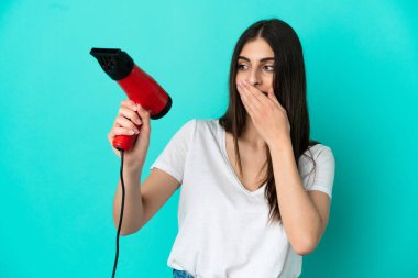 Young caucasian woman holding a hairdryer isolated on blue background with surprise and shocked facial expression